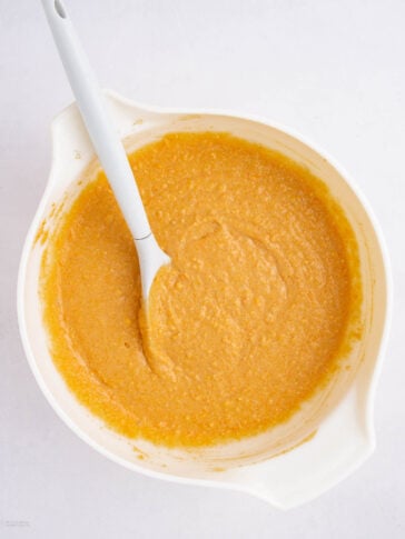 A white mixing bowl filled with orange pumpkin batter sits on a white surface, with a white spatula partially submerged and resting on the bowl’s edge.