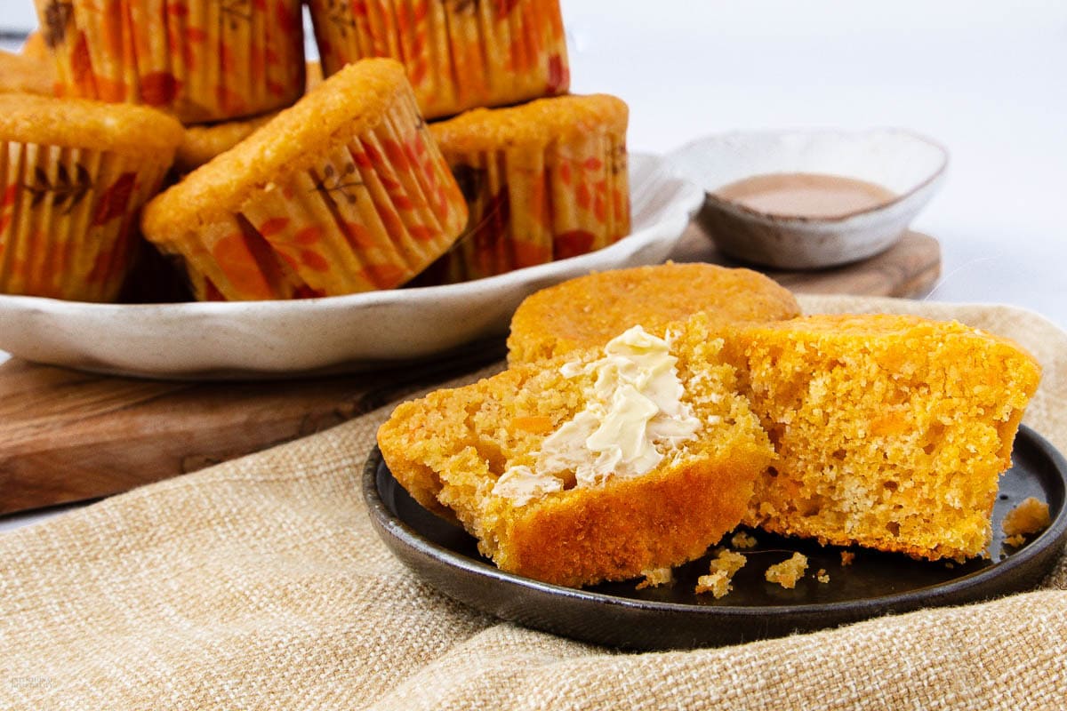 A close-up of sliced cornbread muffins on a plate, one half spread with butter. More muffins are stacked in the background, with a bowl of sauce nearby, all set on a light-colored cloth and wooden board.