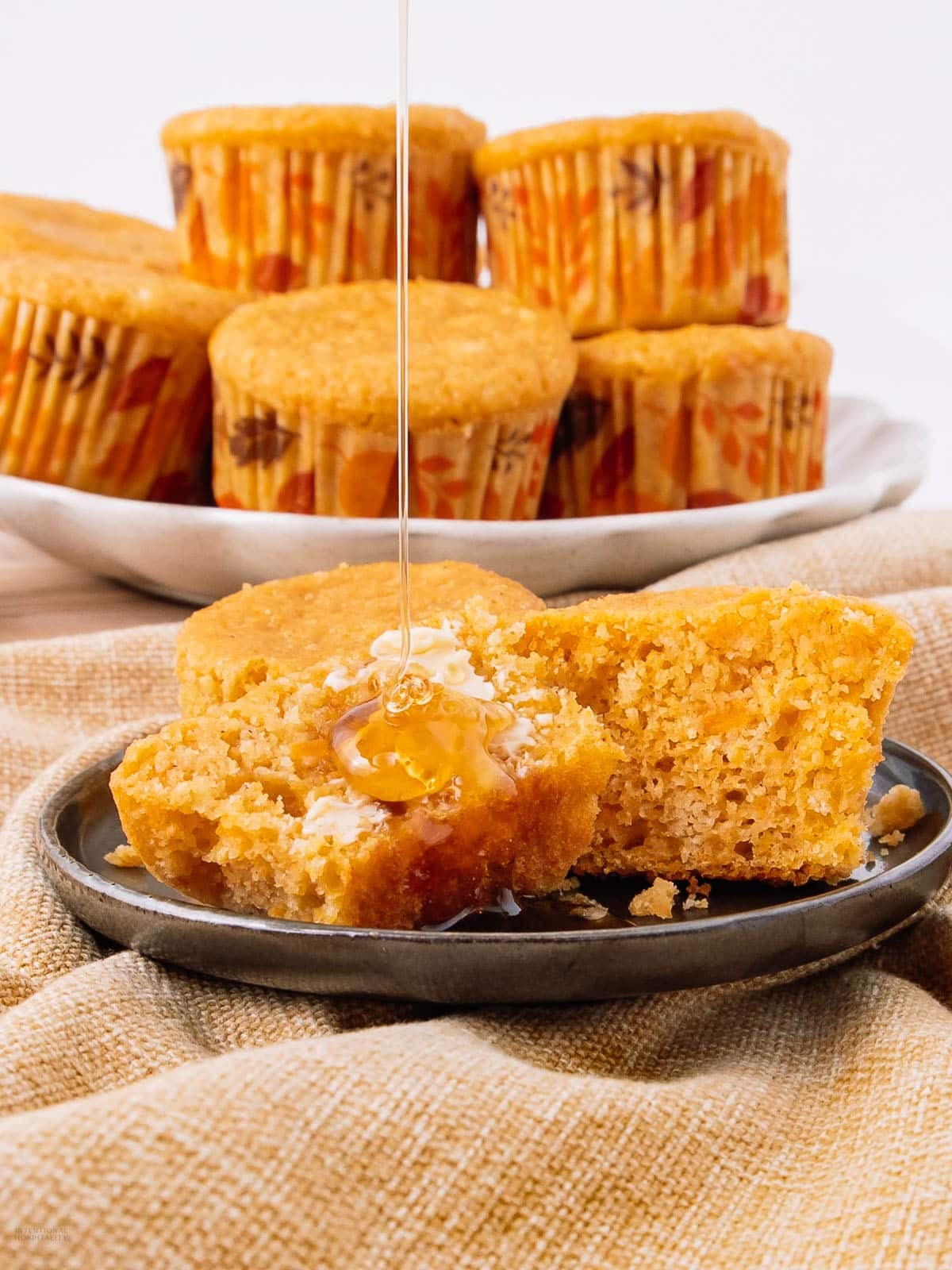 A split cornbread muffin on a plate with butter and honey, surrounded by more muffins in fall-themed wrappers, on a beige cloth.