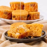 A muffin is split open on a plate with butter and honey drizzling on top. More muffins in autumn-patterned wrappers are stacked on a plate in the background.
