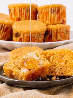 A muffin is split open on a plate with butter and honey drizzling on top. More muffins in autumn-patterned wrappers are stacked on a plate in the background.