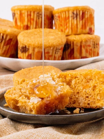 A muffin is split open on a plate with butter and honey drizzling on top. More muffins in autumn-patterned wrappers are stacked on a plate in the background.