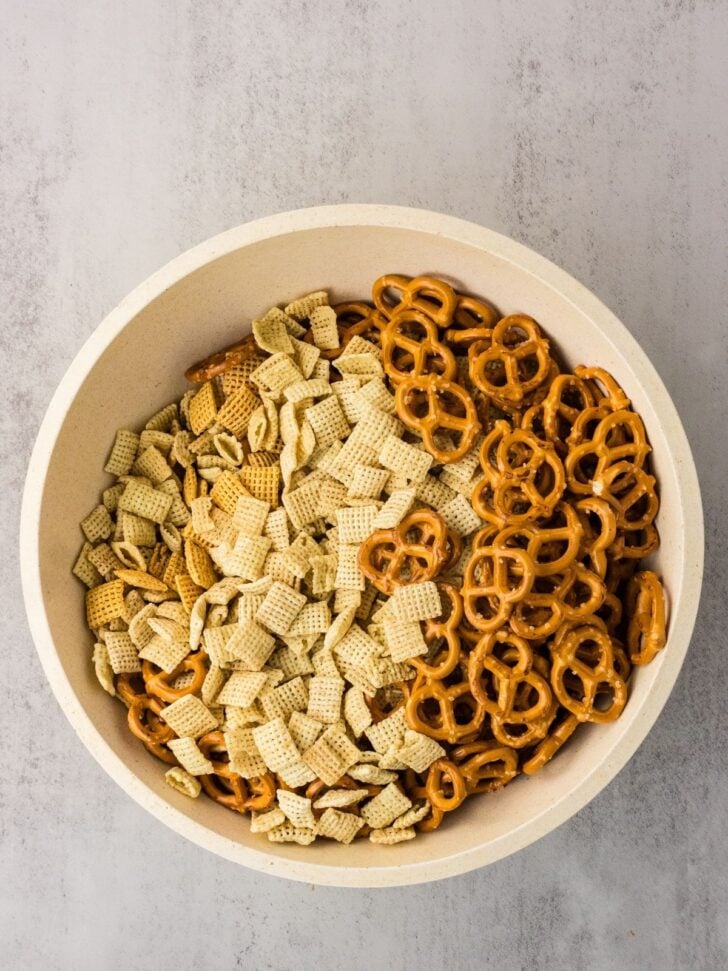 A white bowl filled with mini pretzels and two types of Chex cereal, sitting on a light gray surface.