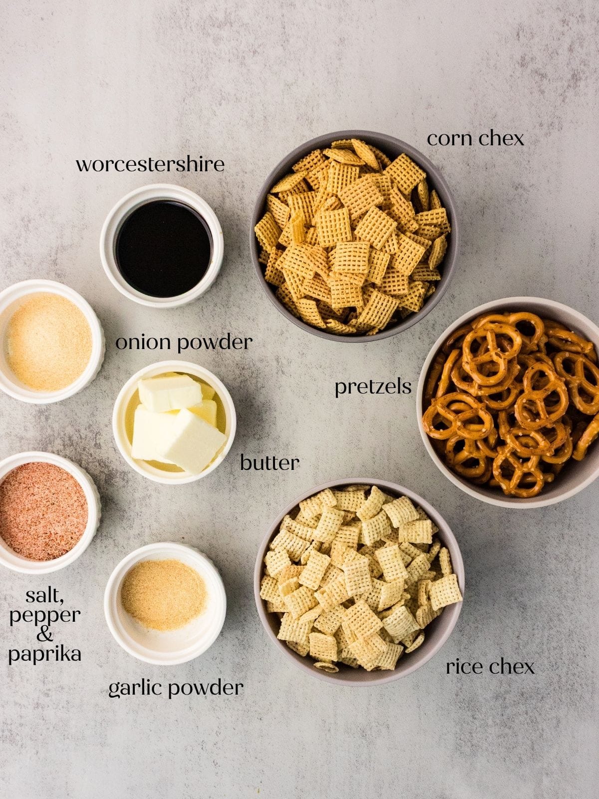 Bowls of corn Chex, rice Chex, pretzels, butter, Worcestershire sauce, garlic powder, onion powder, and a salt, pepper, paprika mix are arranged on a light surface, each labeled with its ingredient name.