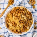 A white bowl filled with a snack mix of pretzels and cereal squares sits on a blue and white checkered cloth, with a wooden spoon and scattered snack pieces nearby.