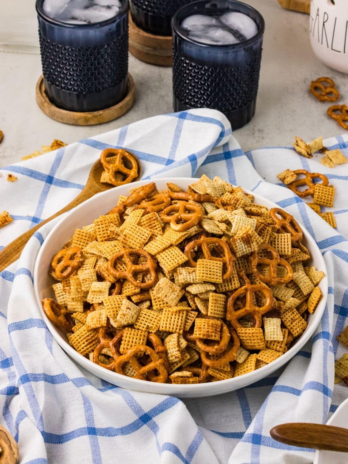 A white bowl filled with Chex mix and pretzels sits on a blue and white checkered cloth, with wooden spoons and glasses of a dark drink nearby.