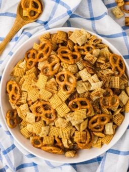 A white bowl filled with a snack mix of pretzels and cereal squares sits on a blue and white checkered cloth, with a wooden spoon and scattered snack pieces nearby.