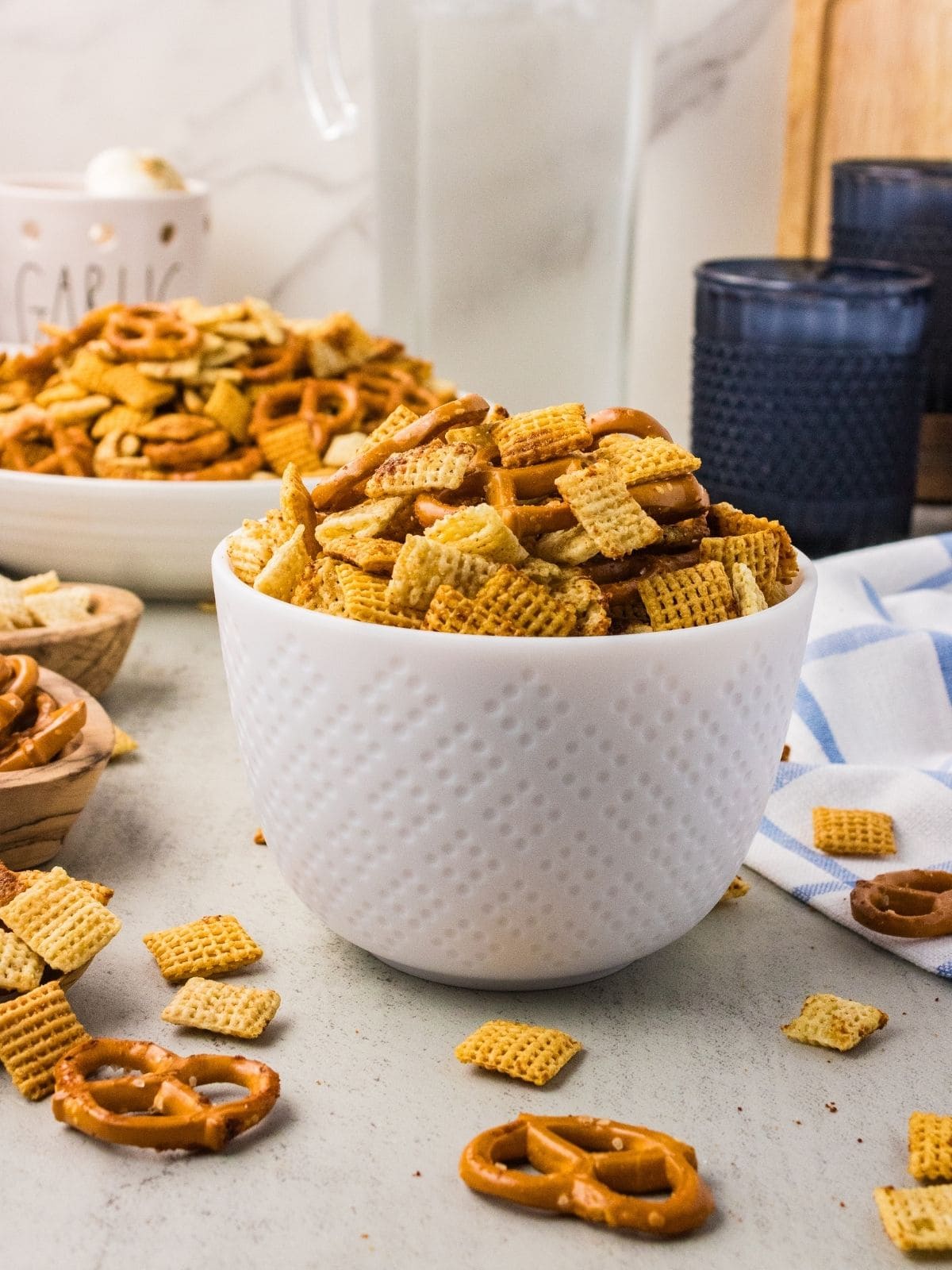 A white bowl filled with snack mix, including pretzels and cereal squares, sits on a light countertop with more snack mix scattered around and a large bowl in the background.