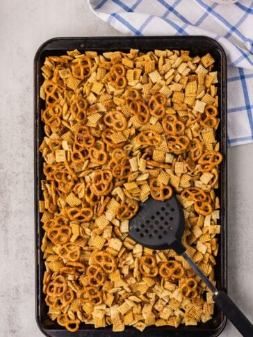 A baking sheet filled with Chex mix and pretzels, stirred with a black slotted spatula. A blue and white checkered towel is partially visible in the background.