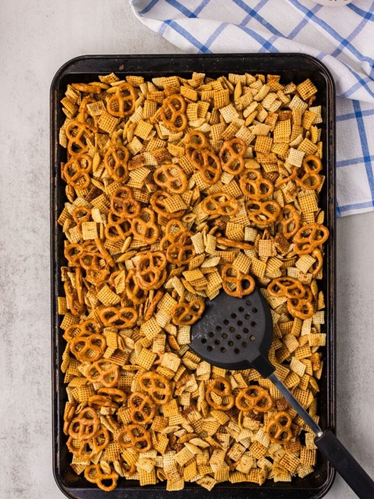 A baking sheet filled with Chex mix and pretzels, stirred with a black slotted spatula. A blue and white checkered towel is partially visible in the background.