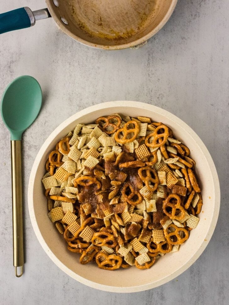 A bowl filled with Chex cereal, pretzels, and seasoning sits on a light surface. Nearby are a green spoon and an empty saucepan with residue visible inside.