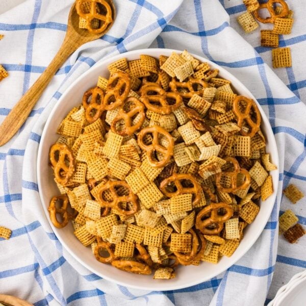 A white bowl filled with a snack mix of pretzels and cereal squares sits on a blue and white checkered cloth, with a wooden spoon and scattered snack pieces nearby.