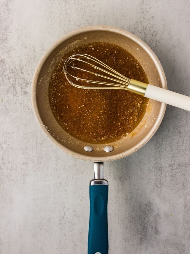 A saucepan with a blue handle contains a brown liquid and a metal whisk with a white handle, placed on a light gray textured surface.