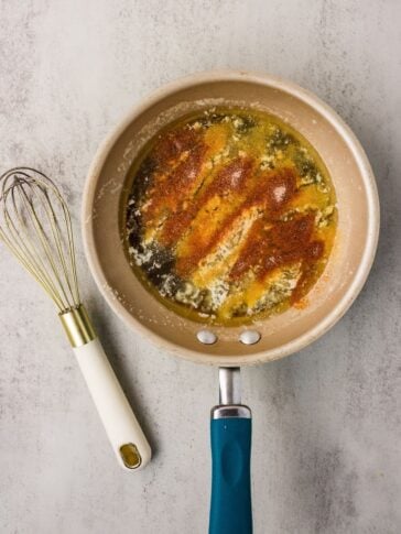 A beige frying pan with melted butter and spices sits on a gray surface next to a metal whisk with a white handle.