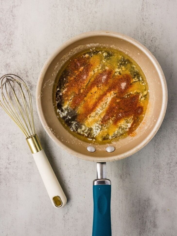 A beige frying pan with melted butter and spices sits on a gray surface next to a metal whisk with a white handle.