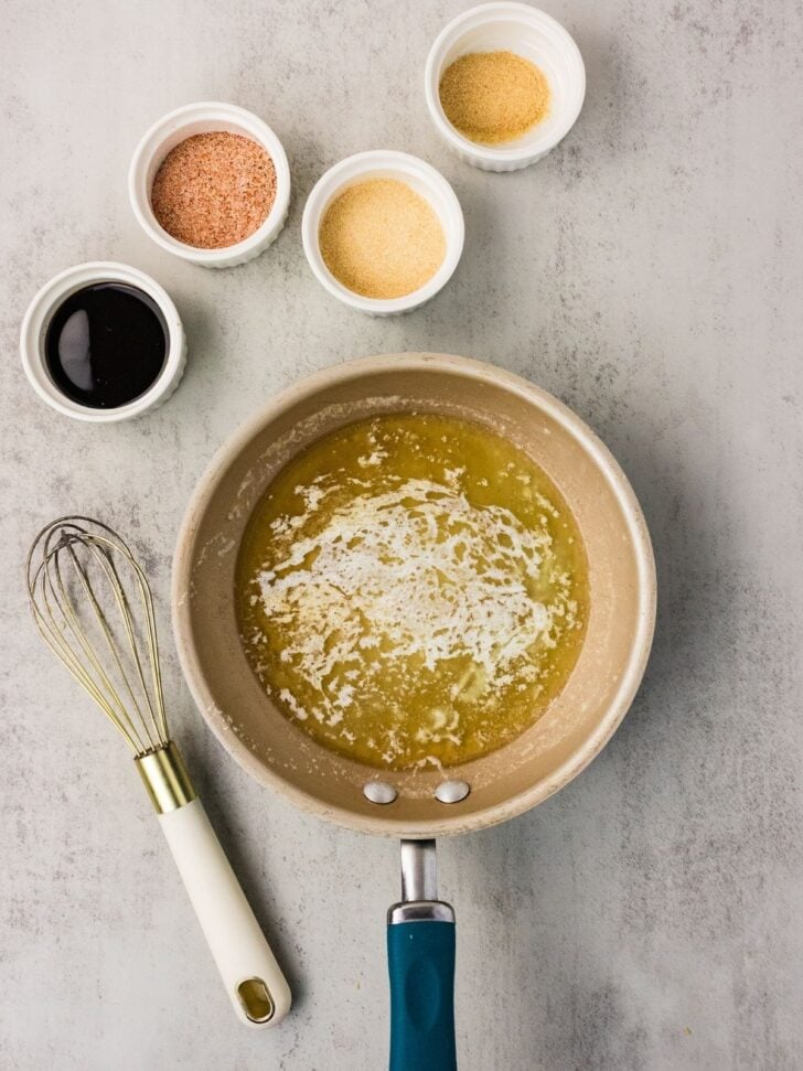 A saucepan with melted butter sits on a countertop beside a whisk. Four small bowls containing spices and a dark liquid are arranged above the pan.