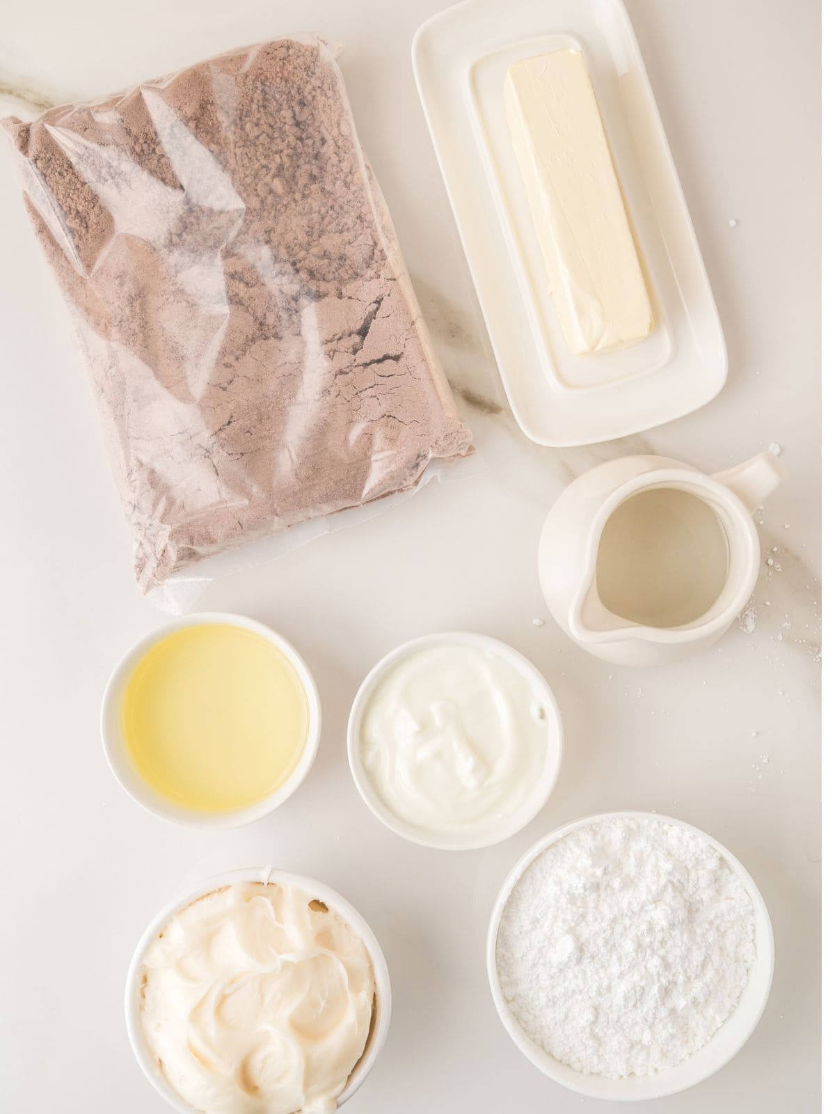 Baking ingredients on a white surface: a bag of cocoa mix, a stick of butter, a small jug of milk, oil, yogurt, a bowl of whipped cream, and a bowl of powdered sugar.