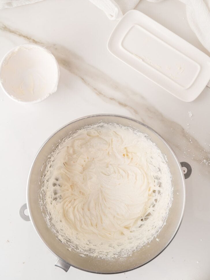 A mixing bowl filled with whipped cream cheese on a marble countertop, with a small empty bowl and a butter dish nearby.