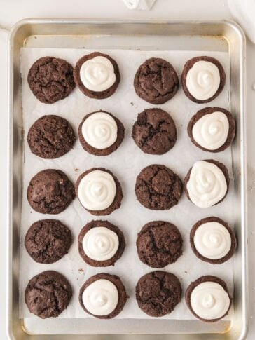 A baking tray with rows of chocolate cookies, some topped with swirled white frosting and others plain, arranged neatly on parchment paper.