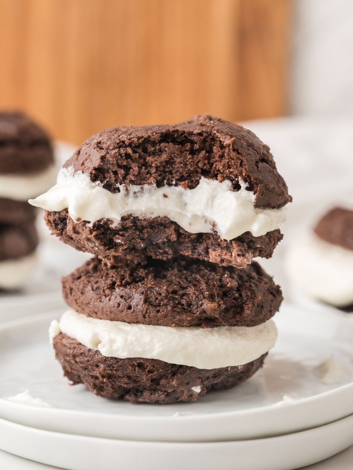 Two chocolate whoopie pies with white cream filling are stacked on a white plate. The top whoopie pie has a bite taken out of it, showing the creamy filling inside. Another whoopie pie is blurred in the background.