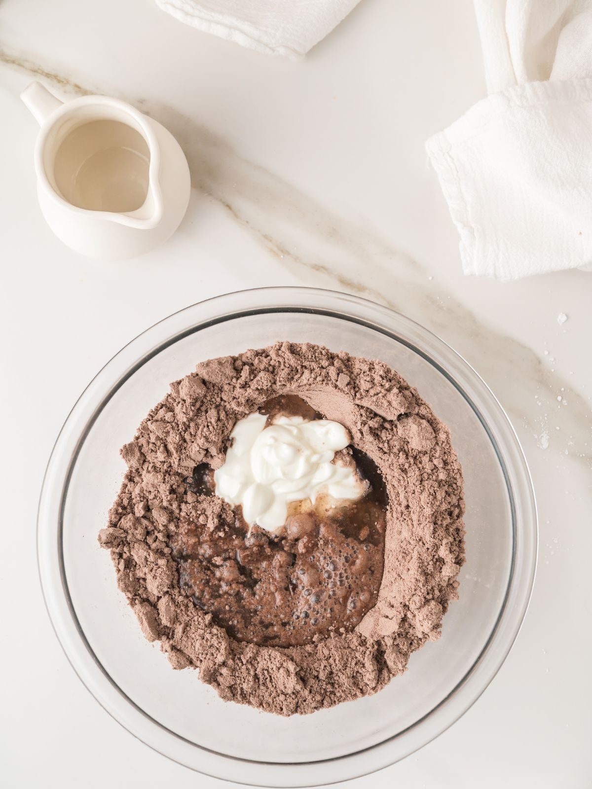 A glass bowl with chocolate cake mix, water, and yogurt on a white counter. A white jug and folded towels are nearby.