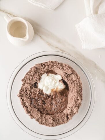 A glass bowl with chocolate cake mix, yogurt, and water on a white countertop, next to a small white pitcher and folded white cloths.