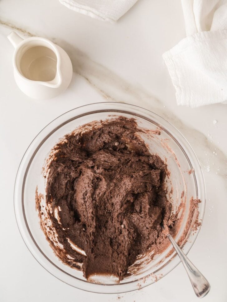 A glass bowl filled with chocolate batter and a spoon sits on a white countertop. A small white pitcher and white cloths are nearby.