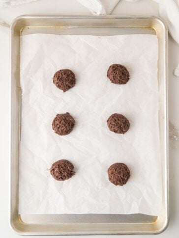 A baking sheet lined with parchment paper holds six evenly spaced chocolate cookies, arranged in two columns of three.