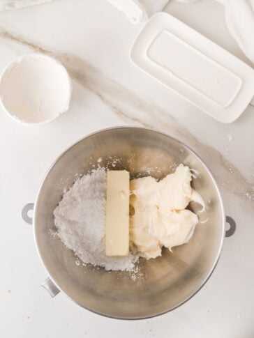 A mixing bowl with powdered sugar, a stick of butter, and cream cheese on a white countertop, next to a butter dish and an empty bowl.