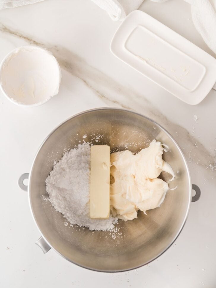 A mixing bowl with powdered sugar, a stick of butter, and cream cheese on a white countertop, next to a butter dish and an empty bowl.