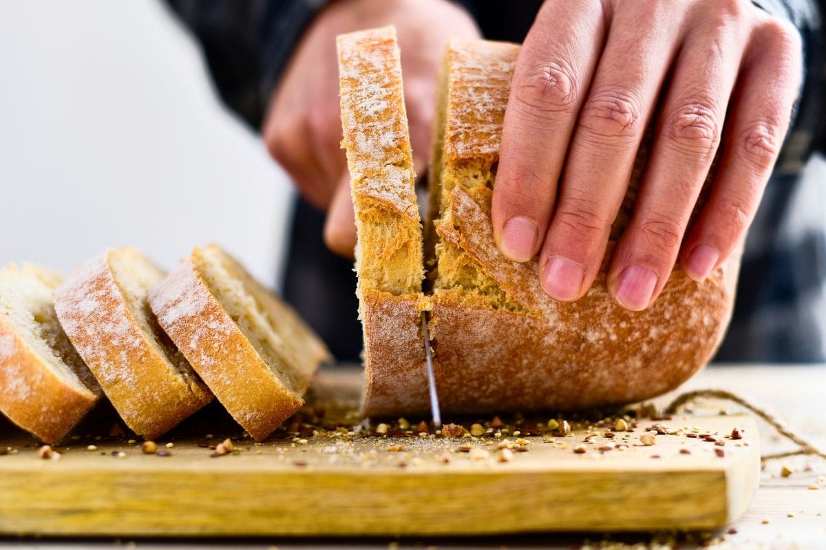 A person slices a round loaf of bread on a wooden cutting board, with several slices already cut and some crumbs scattered around.