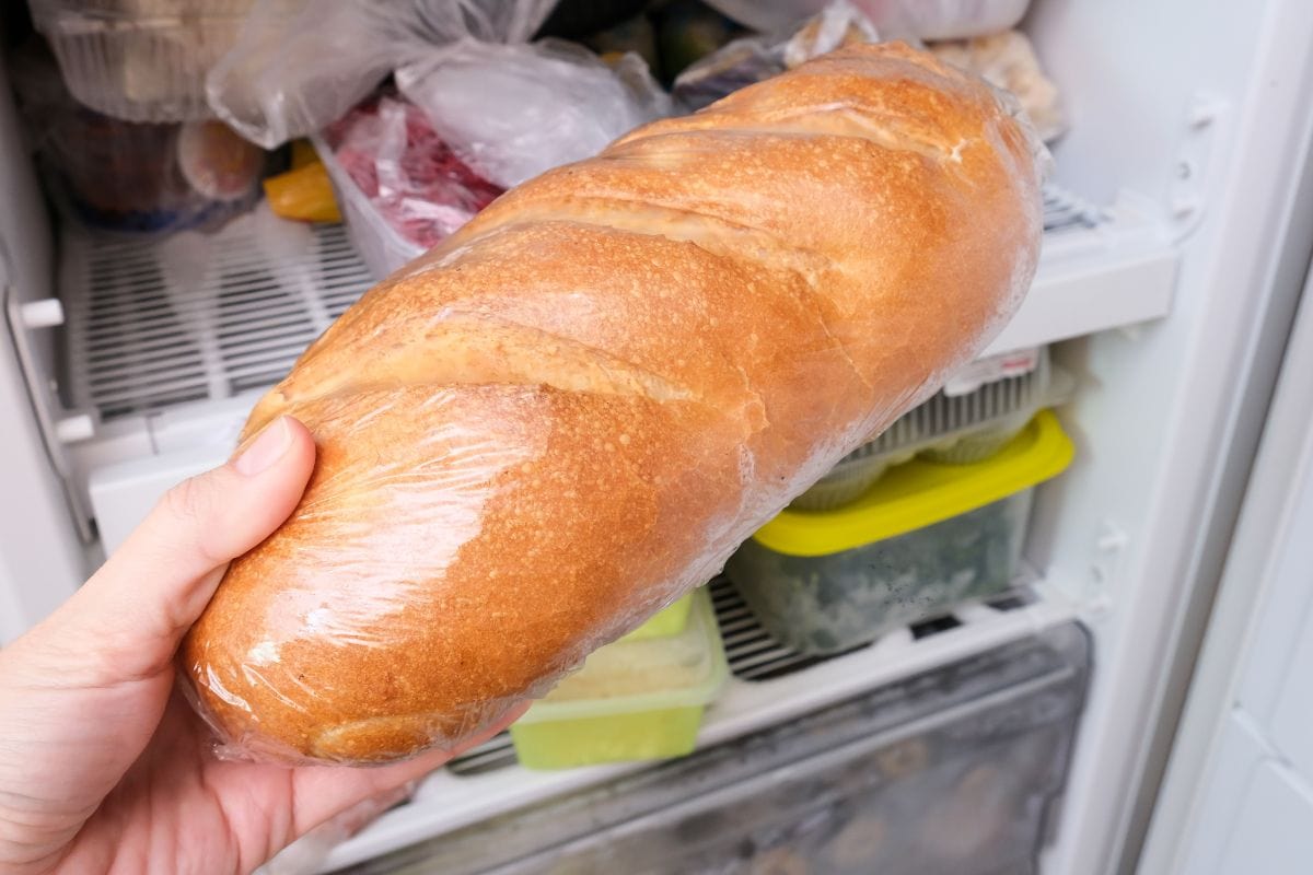 A hand holding a loaf of bread wrapped in plastic wrap, with the loaf being placed into or taken from a freezer filled with various food items.