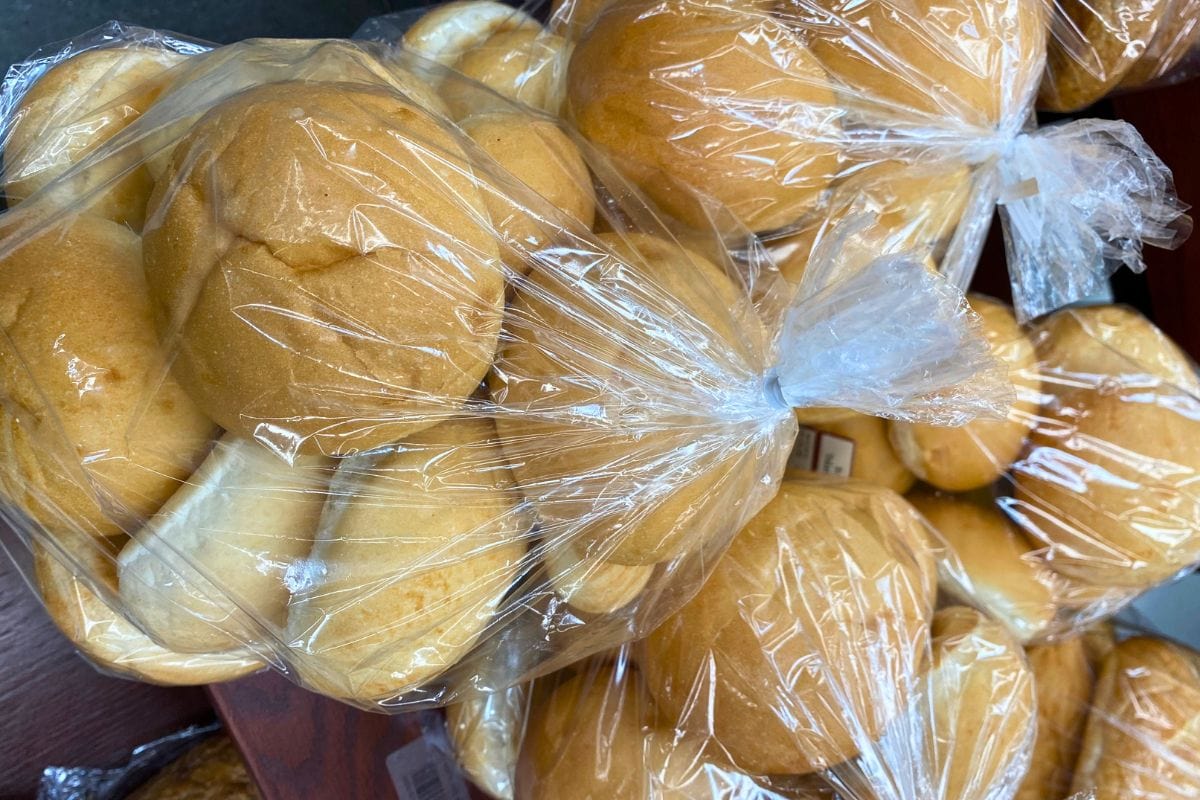 Plastic bags filled with round bread rolls are stacked together on a wooden surface. The rolls are golden brown and the bags are tied at the top.