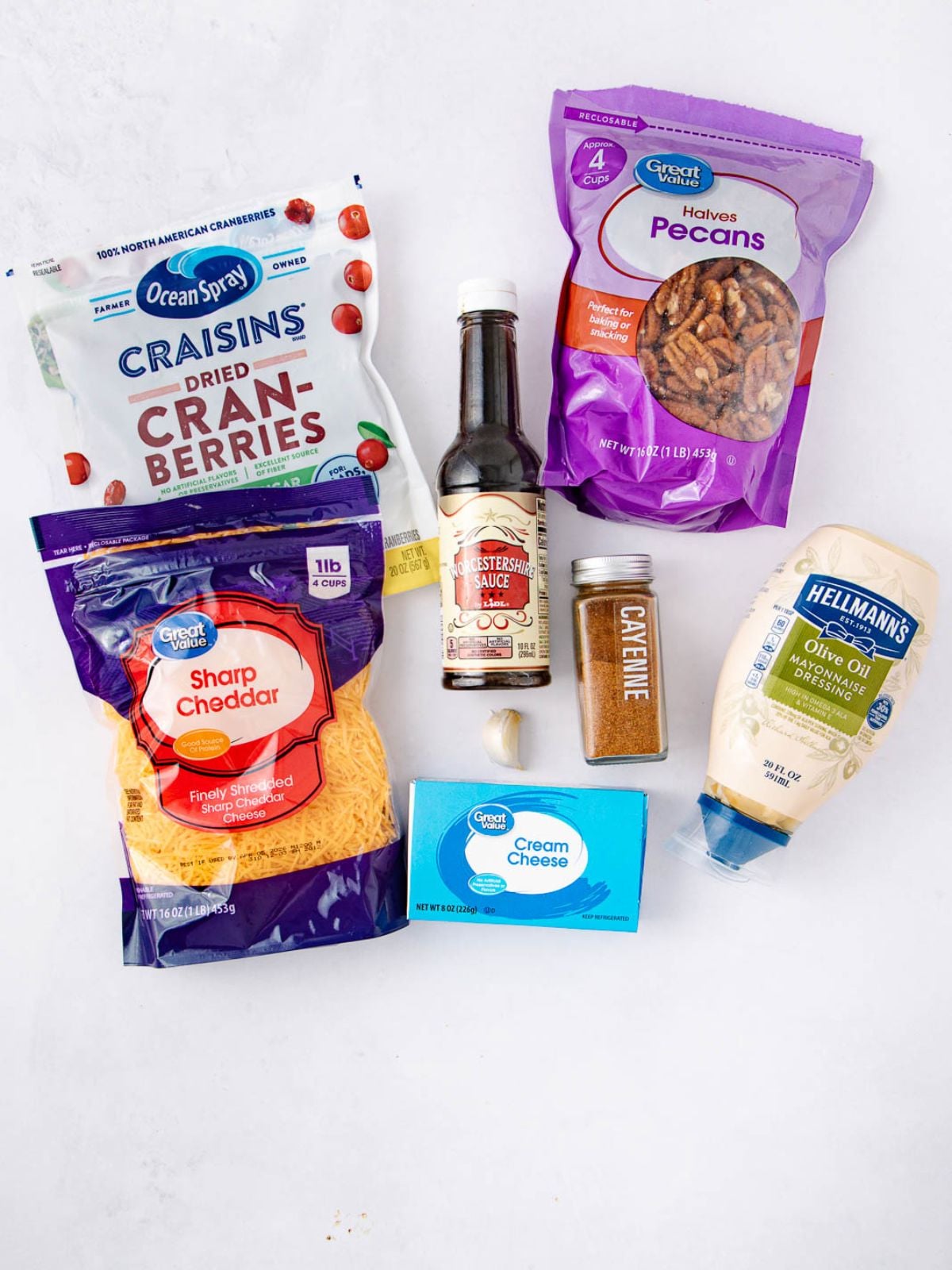 Assorted ingredients on a white surface, including cranberries, pecans, cheddar cheese, cream cheese, mayonnaise, Worcestershire sauce, cayenne pepper, and a garlic clove.