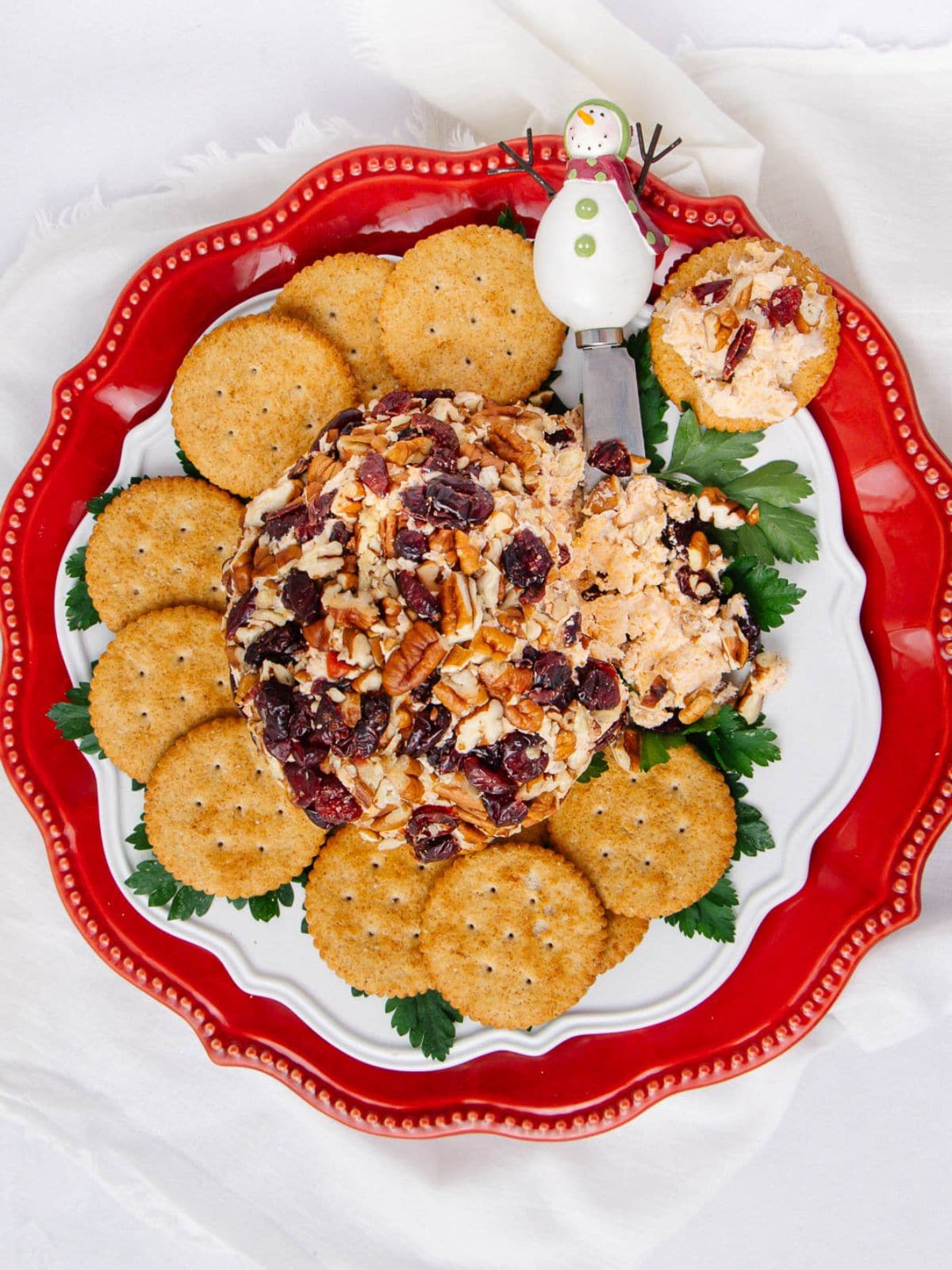 A festive cheese ball topped with nuts and dried cranberries, surrounded by round crackers on a red and white plate, with a snowman spreader on the side.