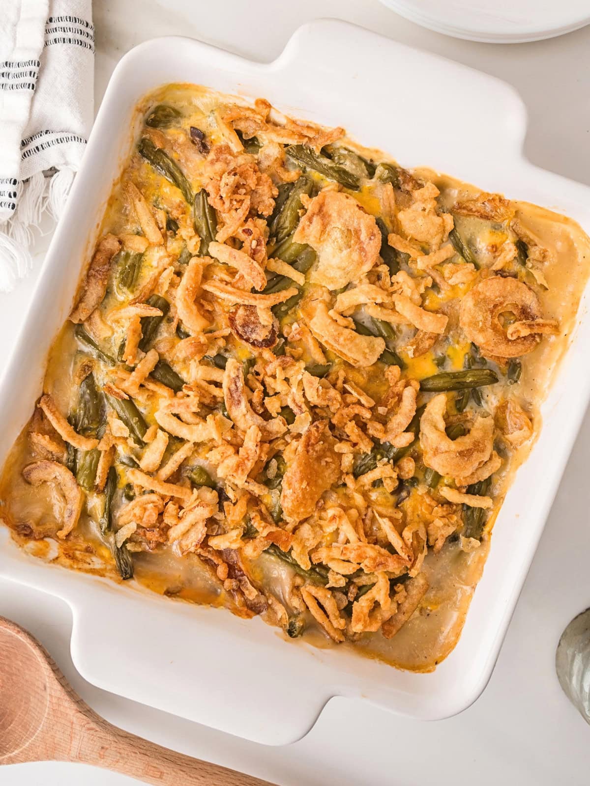 A baked green bean casserole in a white square dish, topped with crispy fried onions. A wooden spoon and part of a striped towel are visible nearby.