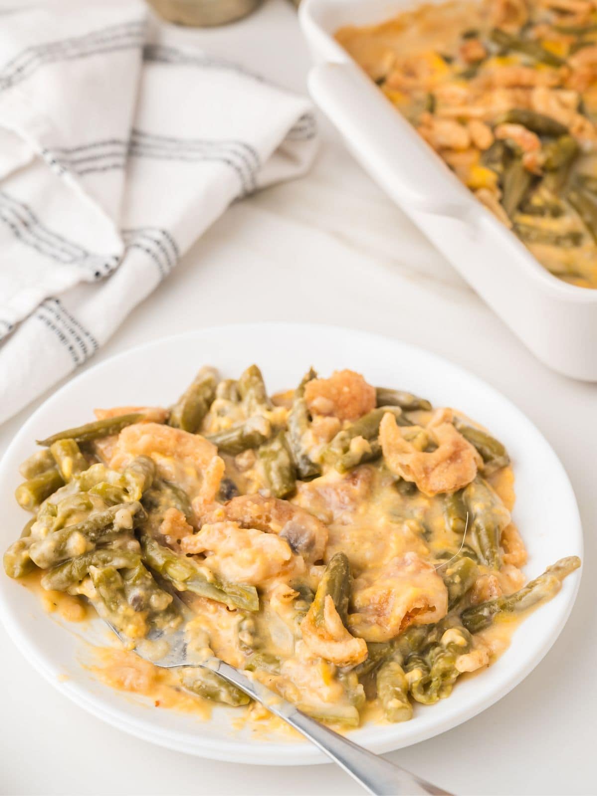 A plate of green bean casserole topped with crispy fried onions sits next to a baking dish of the same dish. A fork rests on the plate, and a striped cloth napkin is nearby.