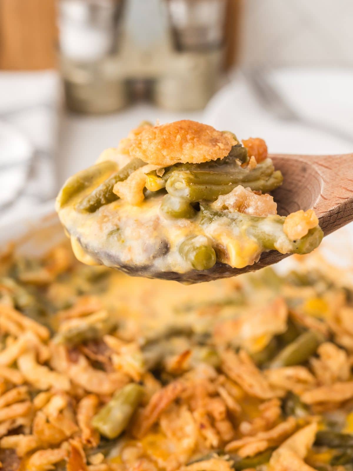 A wooden spoon holds a serving of green bean casserole topped with crispy fried onions, with the rest of the casserole visible in the background.