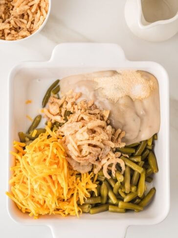 A white dish with green beans, shredded cheddar cheese, crispy fried onions, cream soup, and seasonings, ready to be mixed for a casserole.