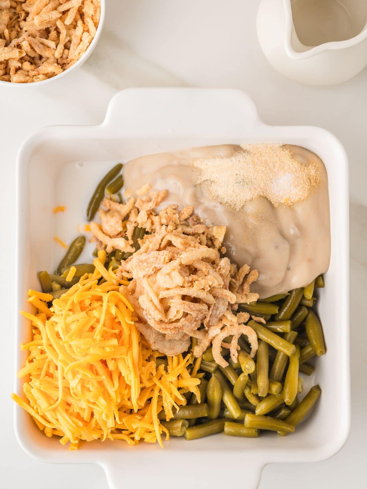 A white dish with green beans, shredded cheddar cheese, crispy fried onions, cream soup, and seasonings, ready to be mixed for a casserole.