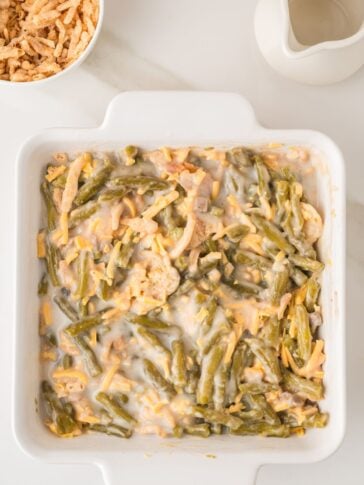 A square white baking dish filled with green bean casserole topped with melted cheese, next to a bowl of crispy onions and a white sauce pitcher on a white surface.