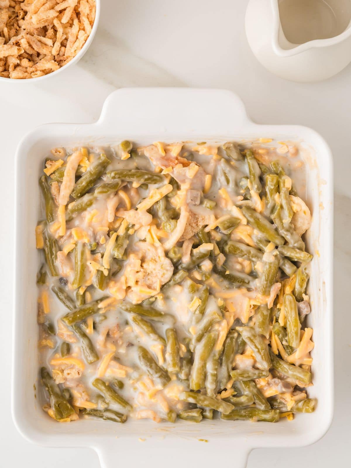 A square white baking dish filled with green bean casserole topped with melted cheese, next to a bowl of crispy onions and a white sauce pitcher on a white surface.