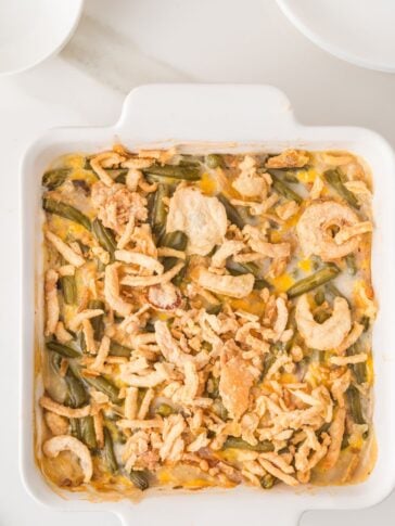 A square white dish filled with green bean casserole topped with crispy fried onions, with plates nearby on a white surface.