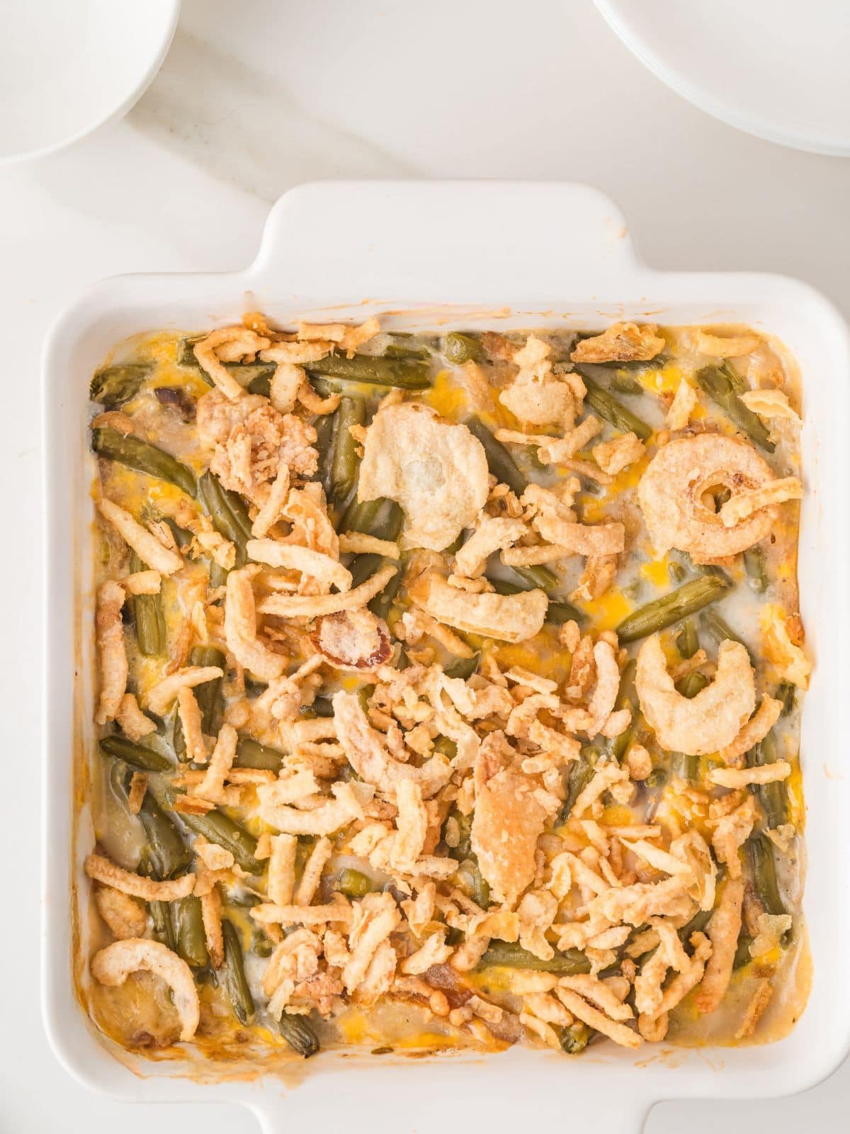 A square white dish filled with green bean casserole topped with crispy fried onions, with plates nearby on a white surface.