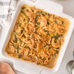 A square white baking dish filled with green bean casserole topped with crispy fried onions, sitting on a white surface near a wooden spoon and a folded cloth napkin.
