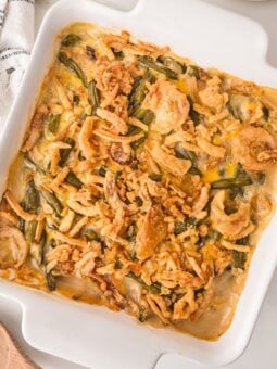 A square white baking dish filled with green bean casserole topped with crispy fried onions, sitting on a white surface near a wooden spoon and a folded cloth napkin.