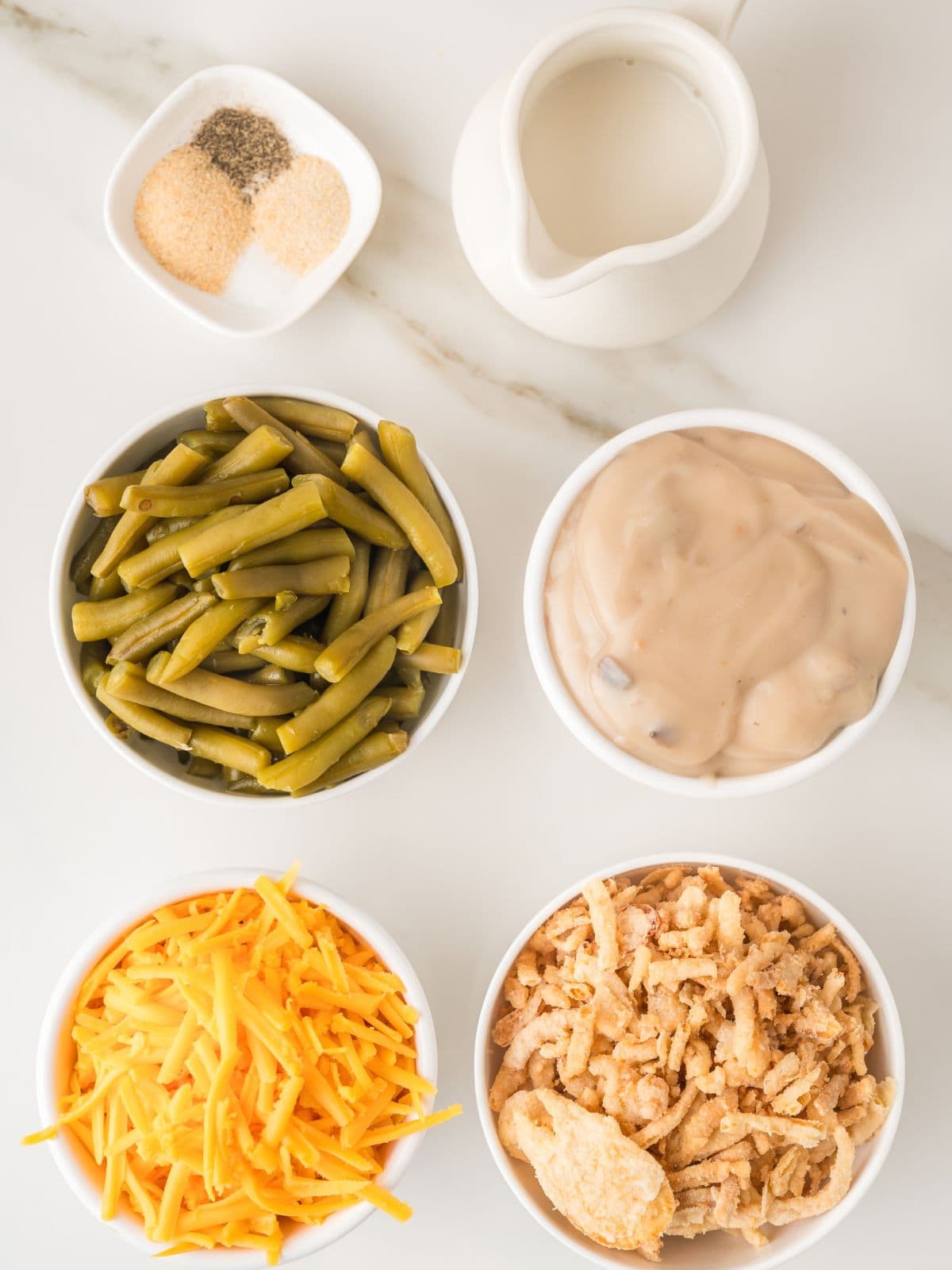 Five bowls with ingredients: shredded cheddar cheese, green beans, crispy fried onions, creamy soup, and seasonings. A small pitcher of milk is also visible on a white surface.