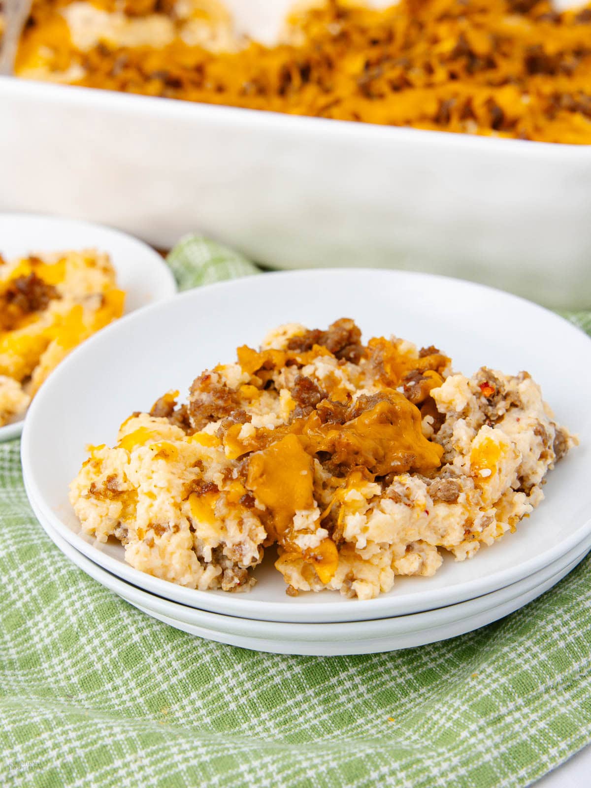 A serving of cheesy breakfast casserole with ground sausage and eggs sits on a white plate, with more casserole visible in a baking dish in the background. The plate rests on a green checkered cloth.