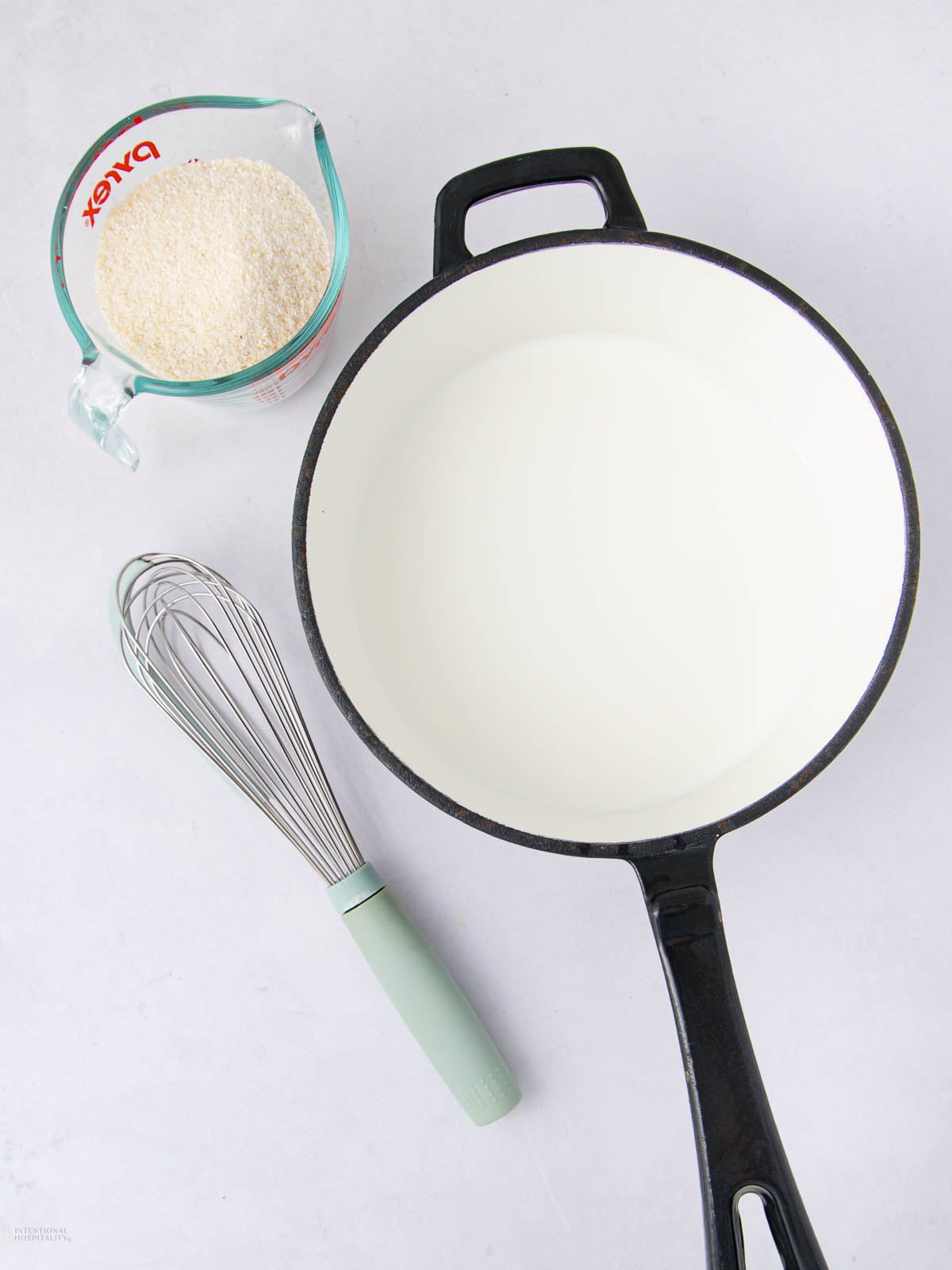 A whisk, a measuring cup filled with white grains, and a white enameled pan with liquid are arranged on a white surface.