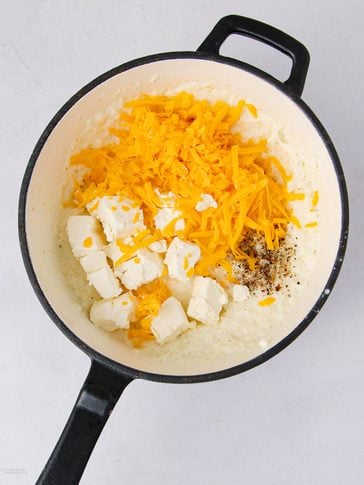 A black pot contains a creamy mixture topped with shredded cheddar cheese, cubed cream cheese, and black pepper, ready to be mixed.